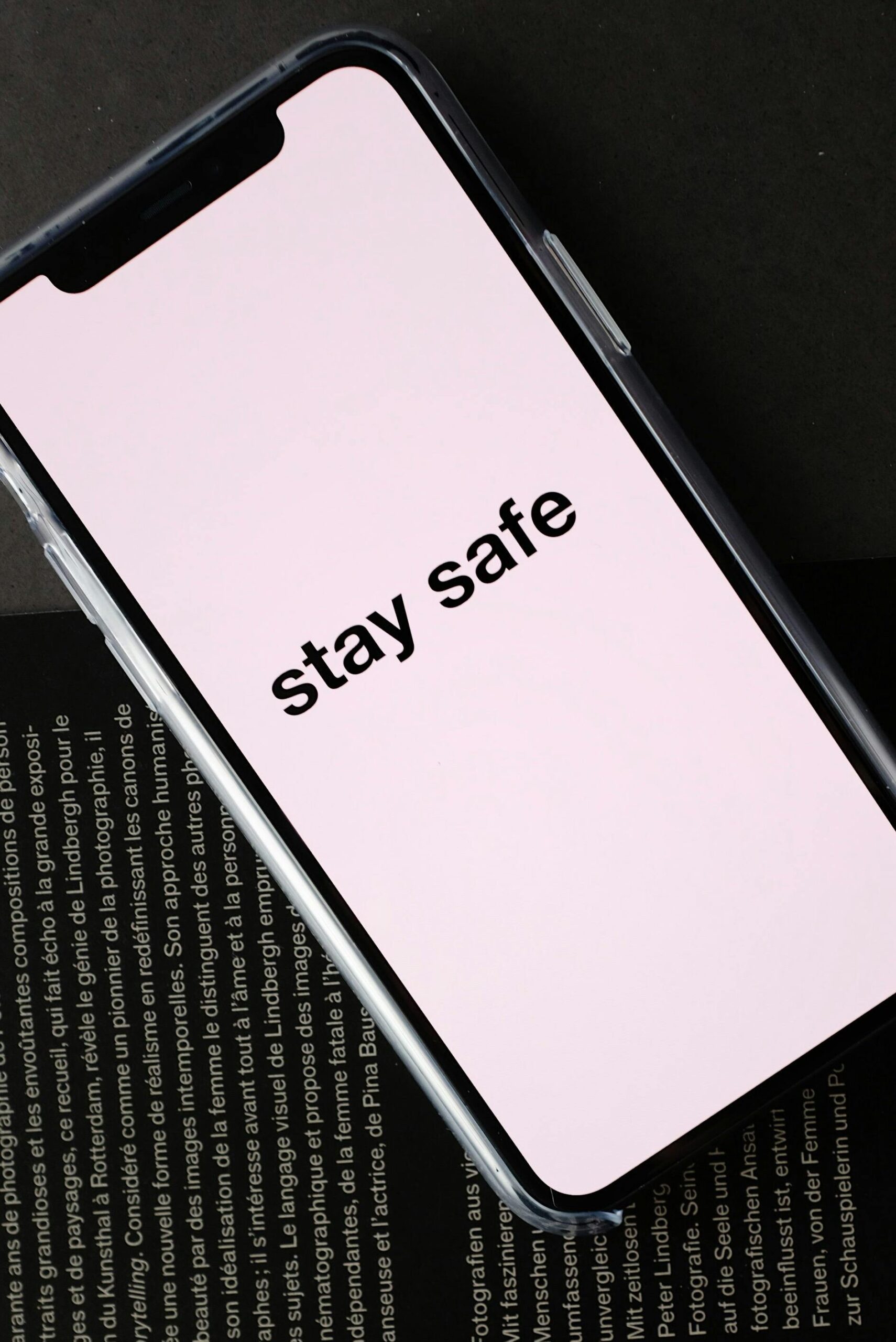 Top view of modern mobile phone with pale pink screen and STAY SAFE inscription placed on open magazine with black page and white letters during COVID 19 pandemic
